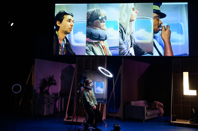 THE VILLAGE: travellers in a plane cabin on screen, with a performer on set below